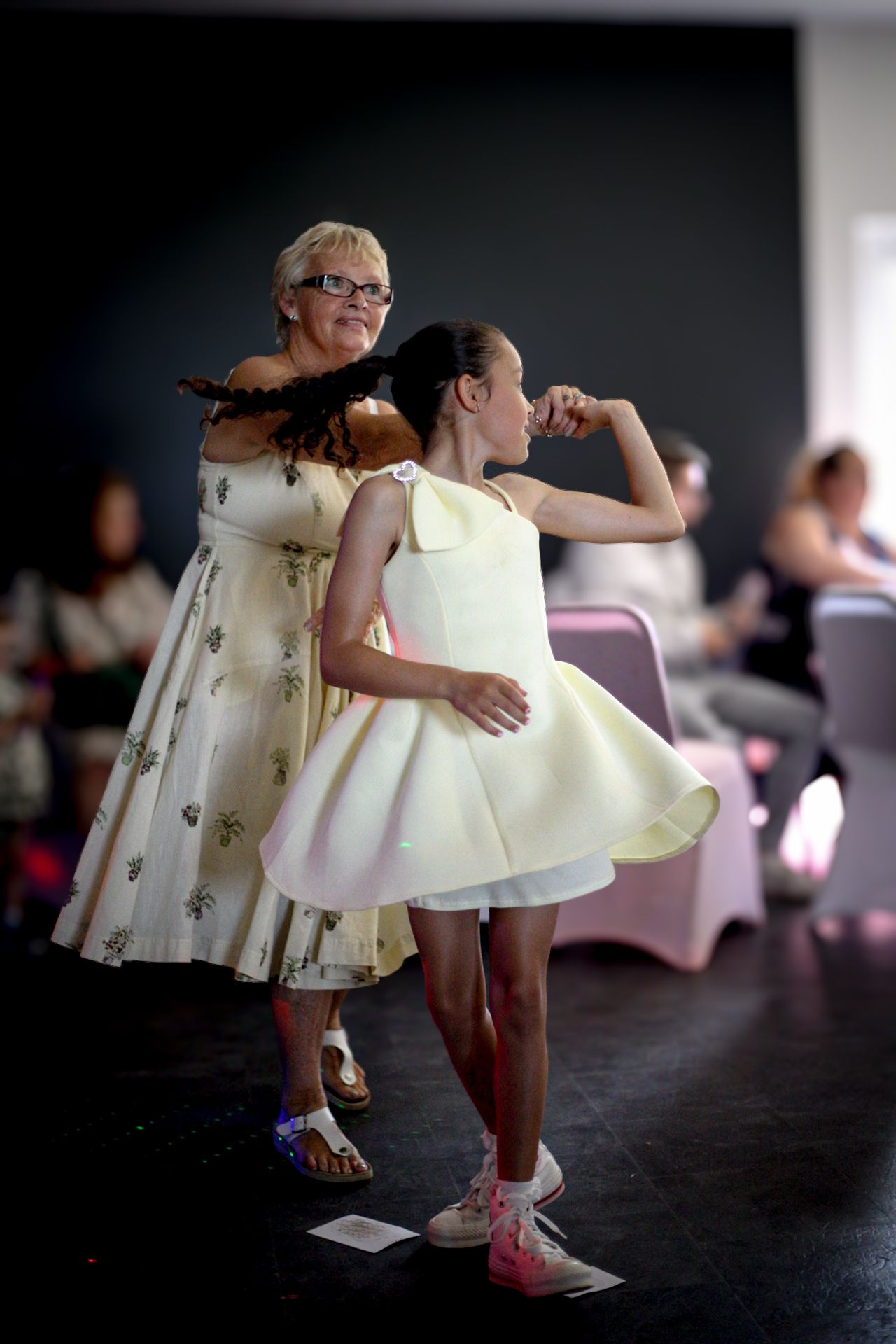 Young dancers in white dresses practice their moves on a dark stage.