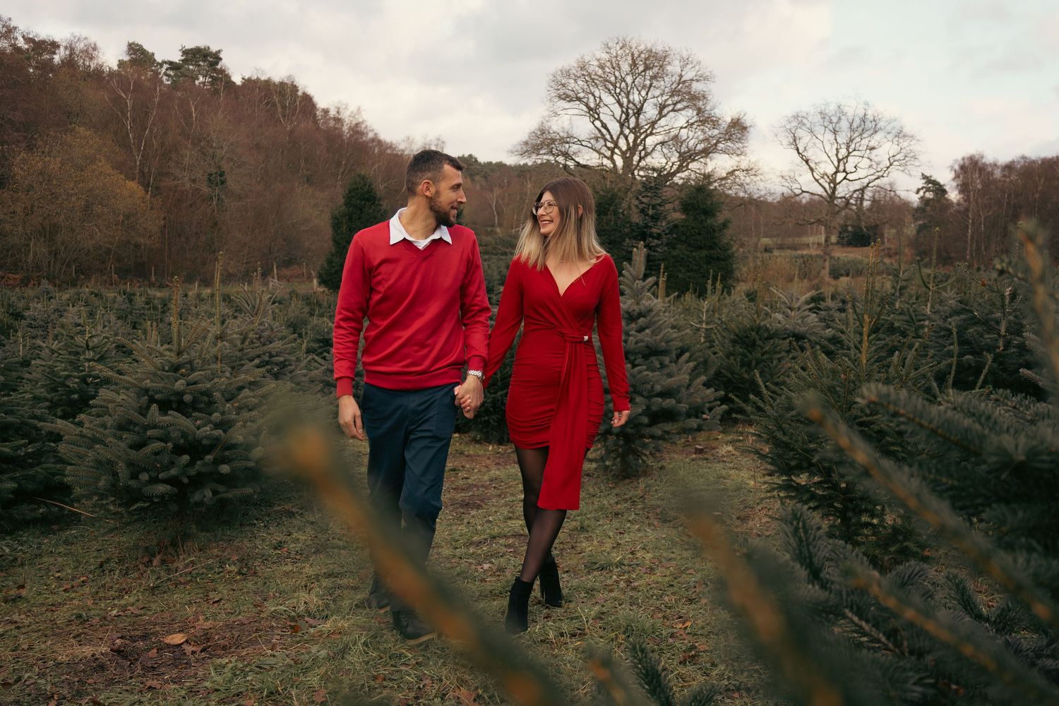Couple holding hands walking through a Christmas tree farm, wearing matching red outfits, surrounded by evergreen trees.