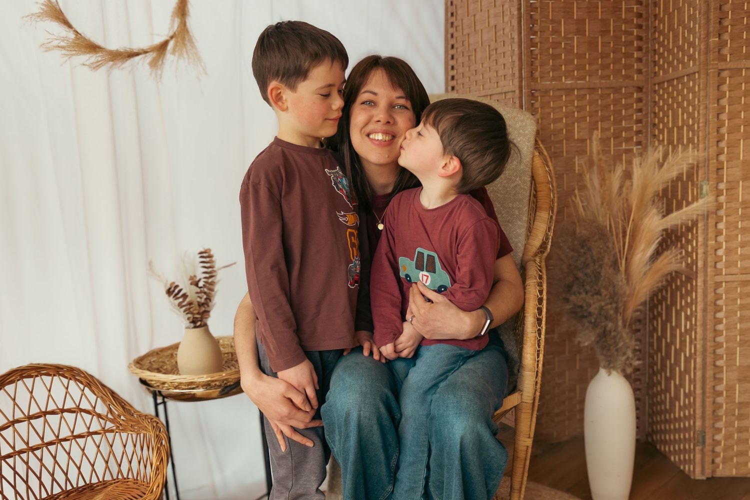 A woman sits in a wicker chair smiling as two young boys in burgundy shirts give her a kiss on the cheek.