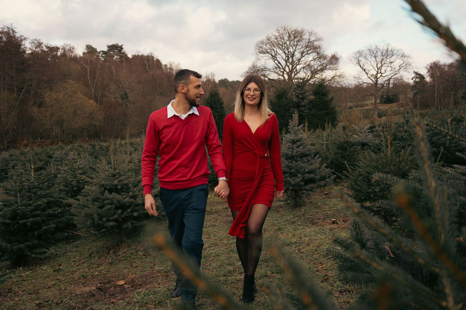 Two people stroll together among evergreen trees on a cloudy autumn day.