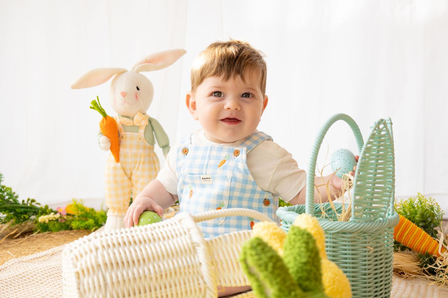 A smiling baby in a gingham outfit surrounded by Easter baskets, toy bunny, and decorative carrots.