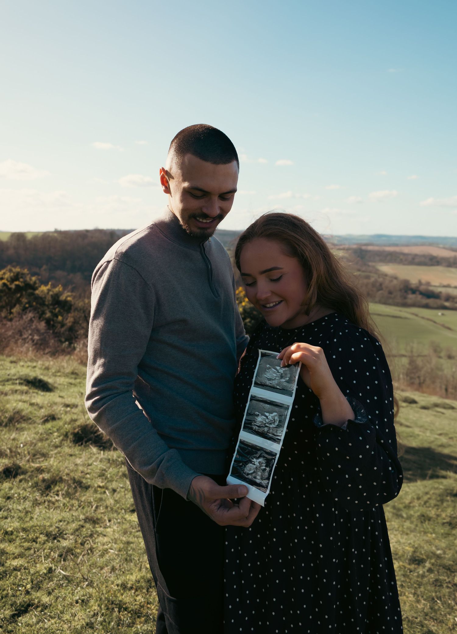 Couple smiling and holding ultrasound scan photos outdoors on a sunny hillside, announcing their pregnancy.