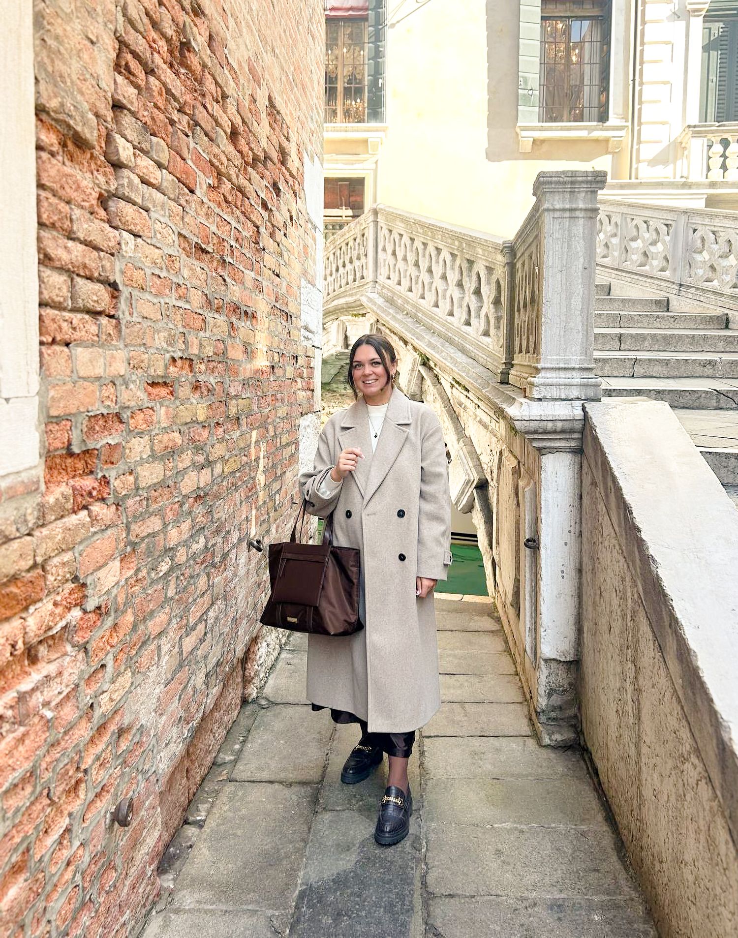 Woman in beige coat with handbag posing on narrow Venice canal walkway near Bridge of Sighs.
