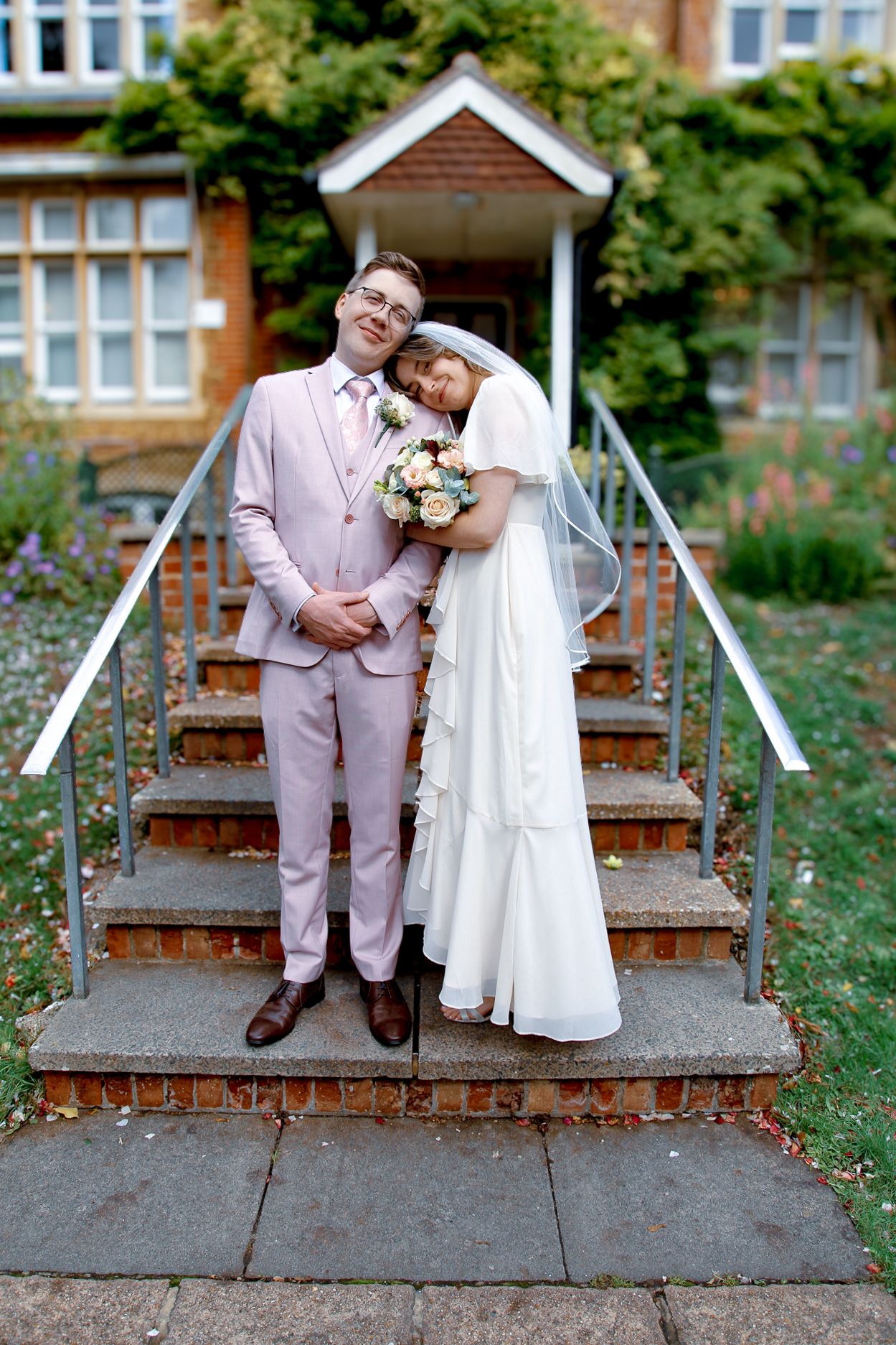 Newlyweds posing on outdoor steps with white gazebo and brick building in background.