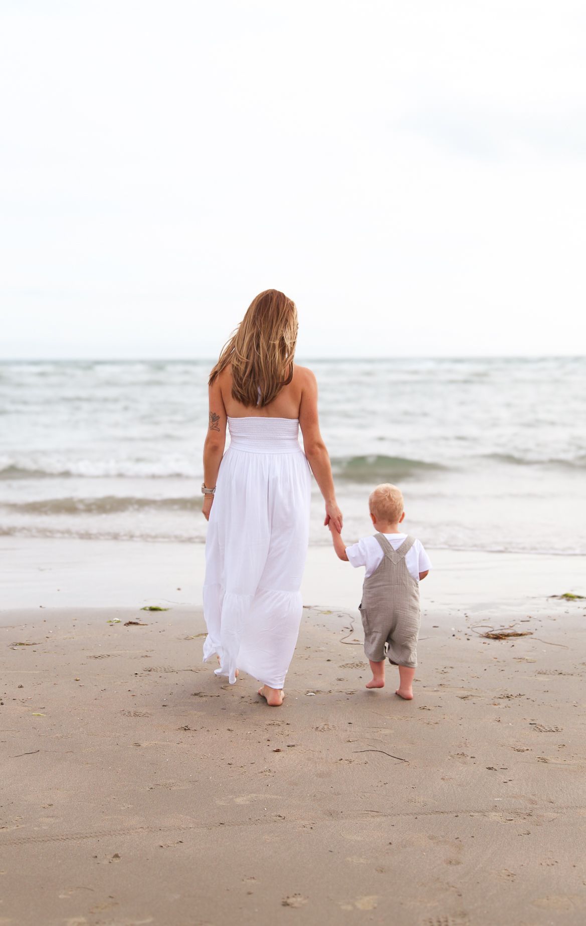 Parent and toddler walking together along empty beach shoreline in white outfits.