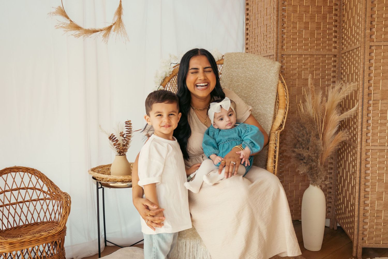 A smiling woman sits in a wicker chair holding a baby in blue, with a young boy standing beside her in a boho studio.
