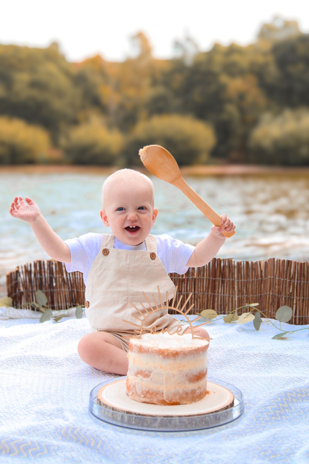 Happy baby sitting outdoors with a wooden spoon celebrating first birthday cake smash near a lake.