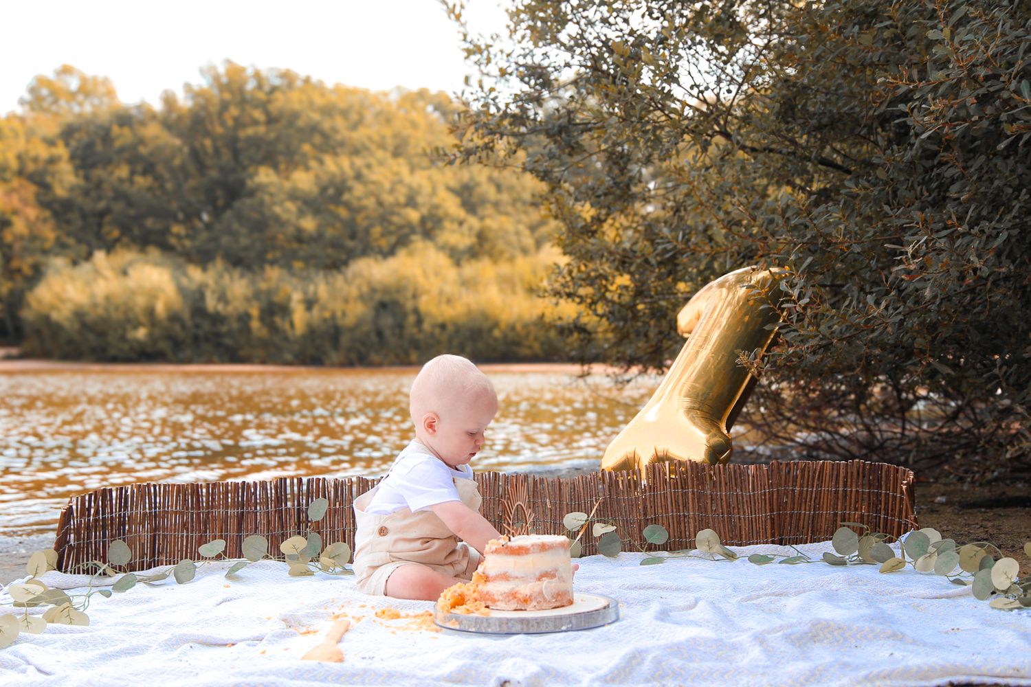 Baby sits on white blanket by lake with gold mermaid tail prop and smash cake during golden hour outdoor photo session.