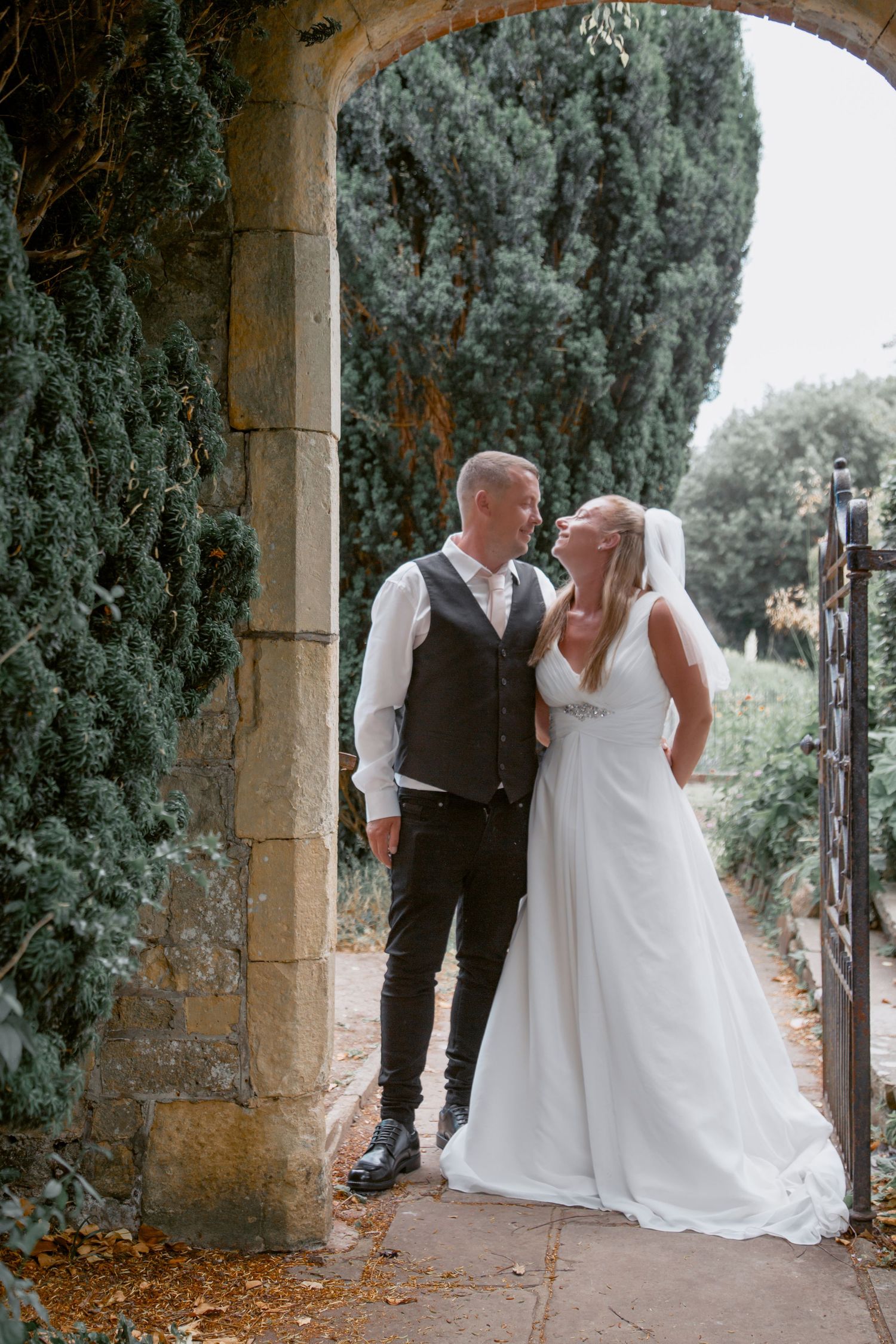 A couple poses romantically under a stone archway surrounded by cypress trees.