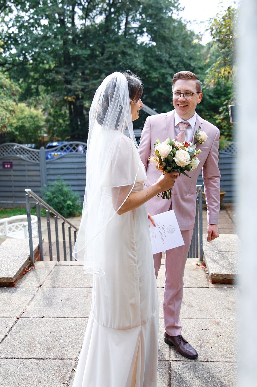 Couple in pink suit and white dress having outdoor wedding photos between white pillars.
