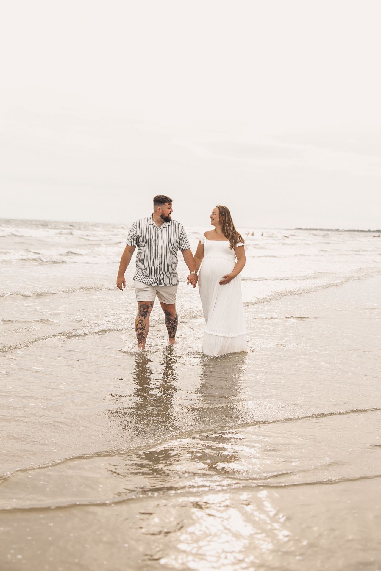 A couple holds hands while walking through gentle ocean waves on a bright beach day.