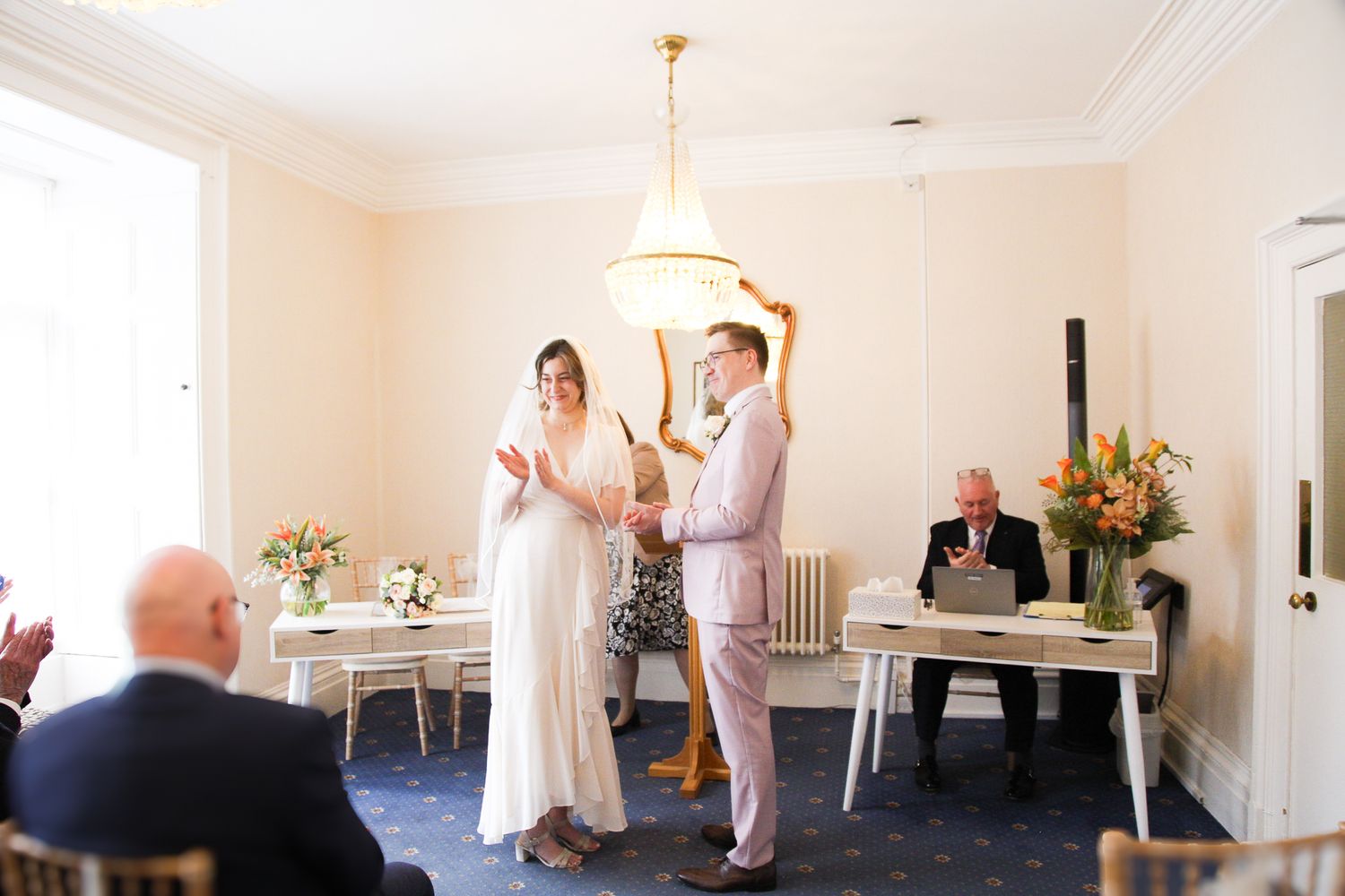 Wedding ceremony in bright indoor space with chandelier and white decor.