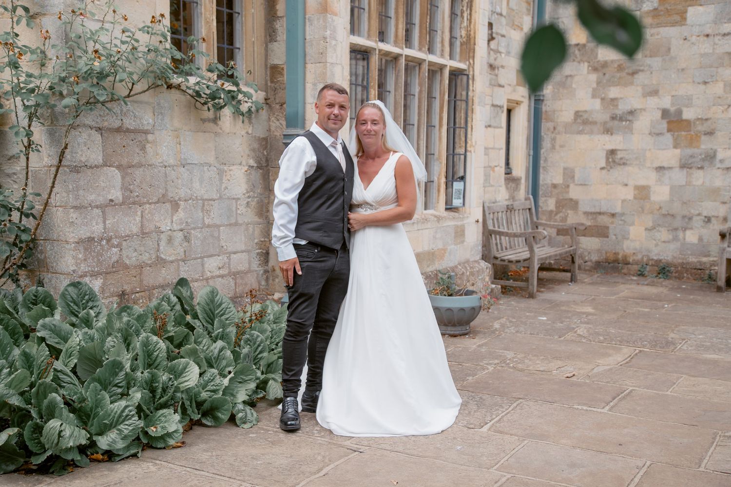 Wedding couple embraces in front of stone building courtyard with lush greenery.