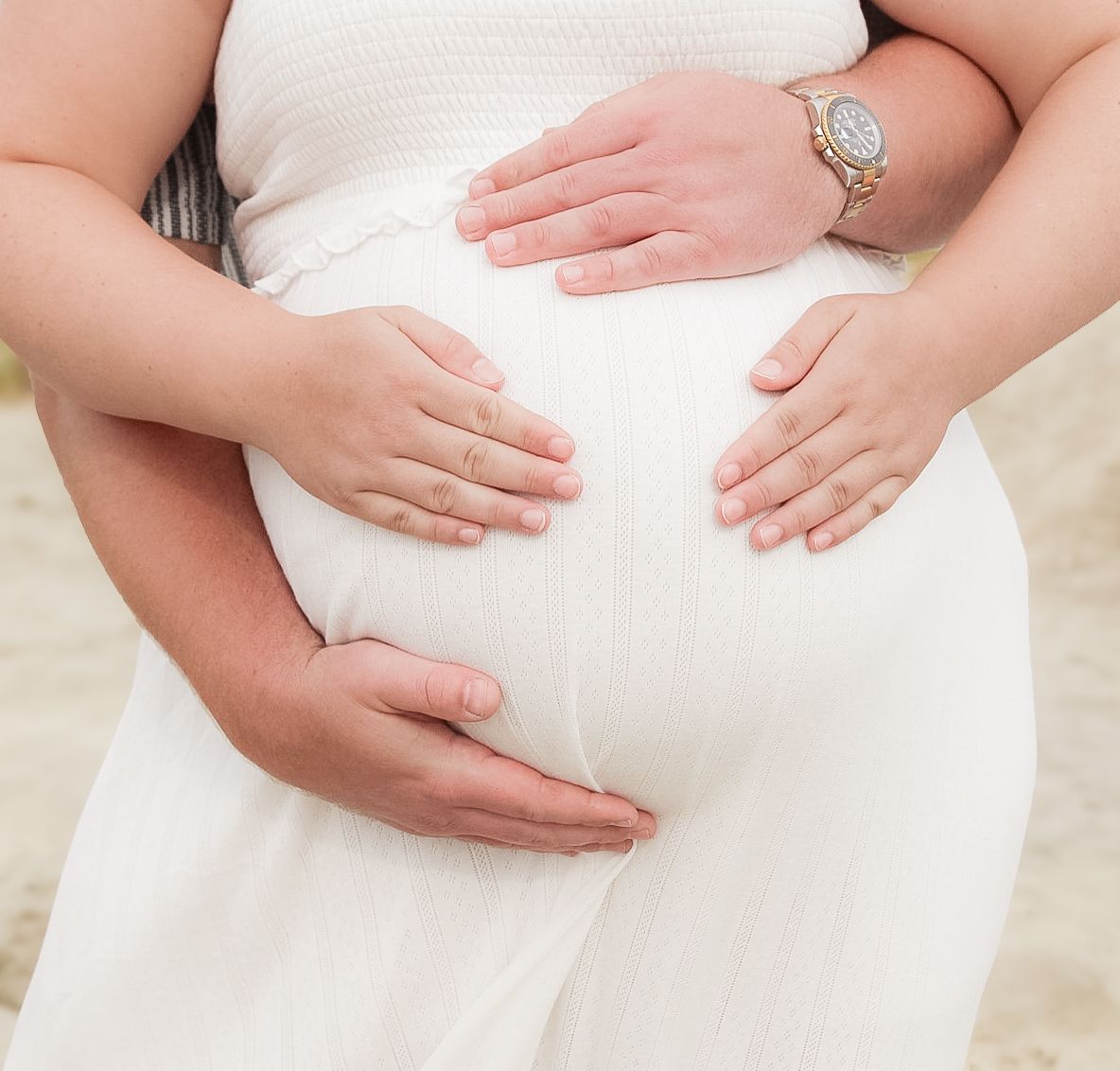 Gentle hands cradle a pregnant belly in white maternity dress.