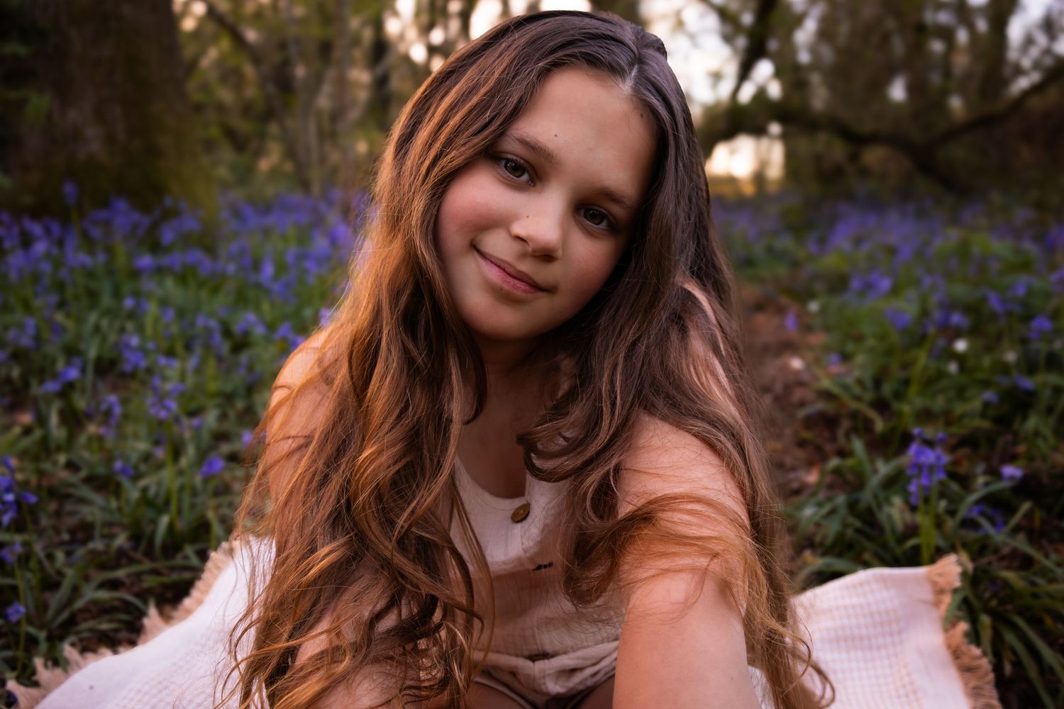 Young girl with long wavy hair sitting among purple bluebells in a woodland garden at dusk.