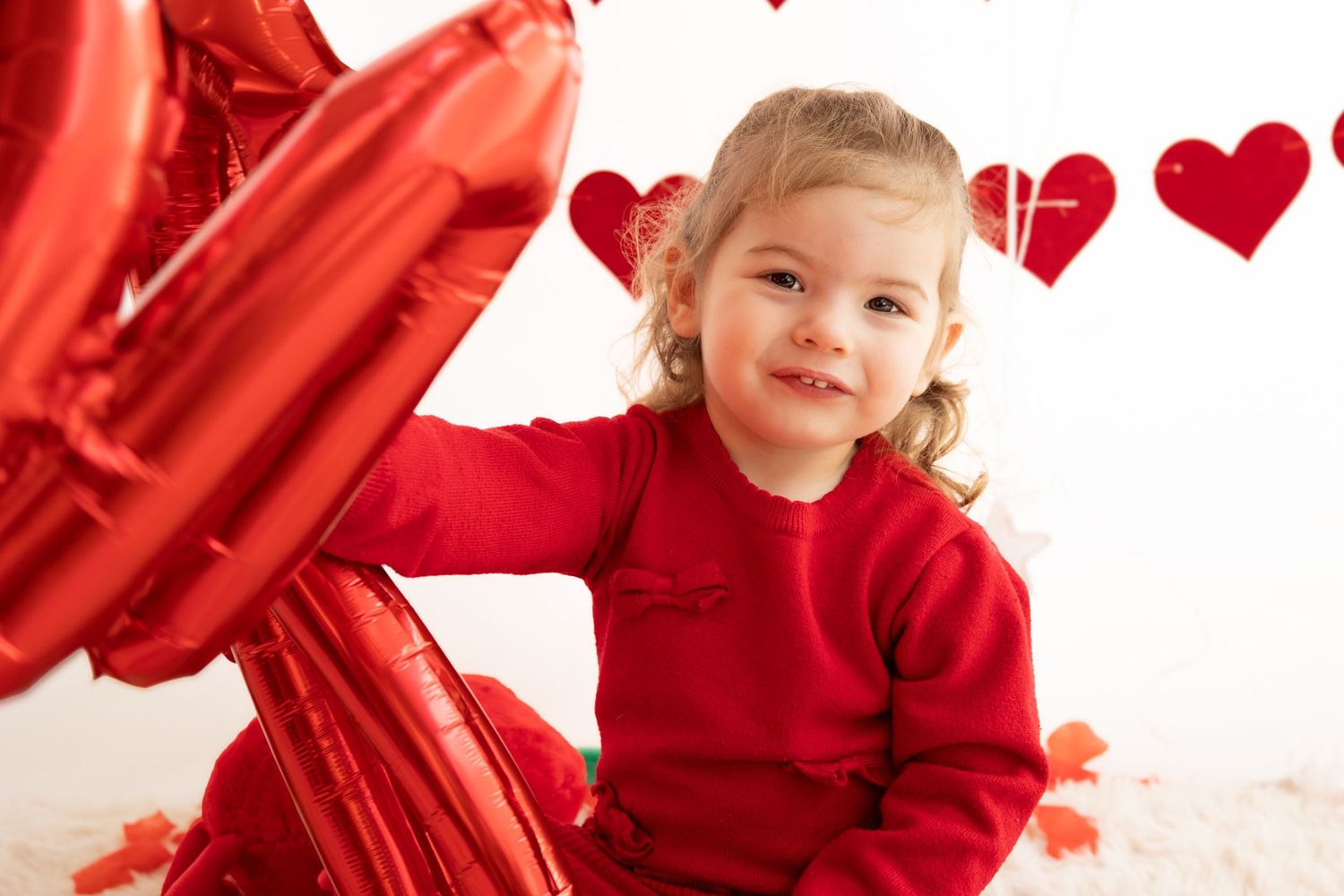 Toddler girl in red outfit smiling with heart balloons and decorations for Valentine's Day photo shoot.