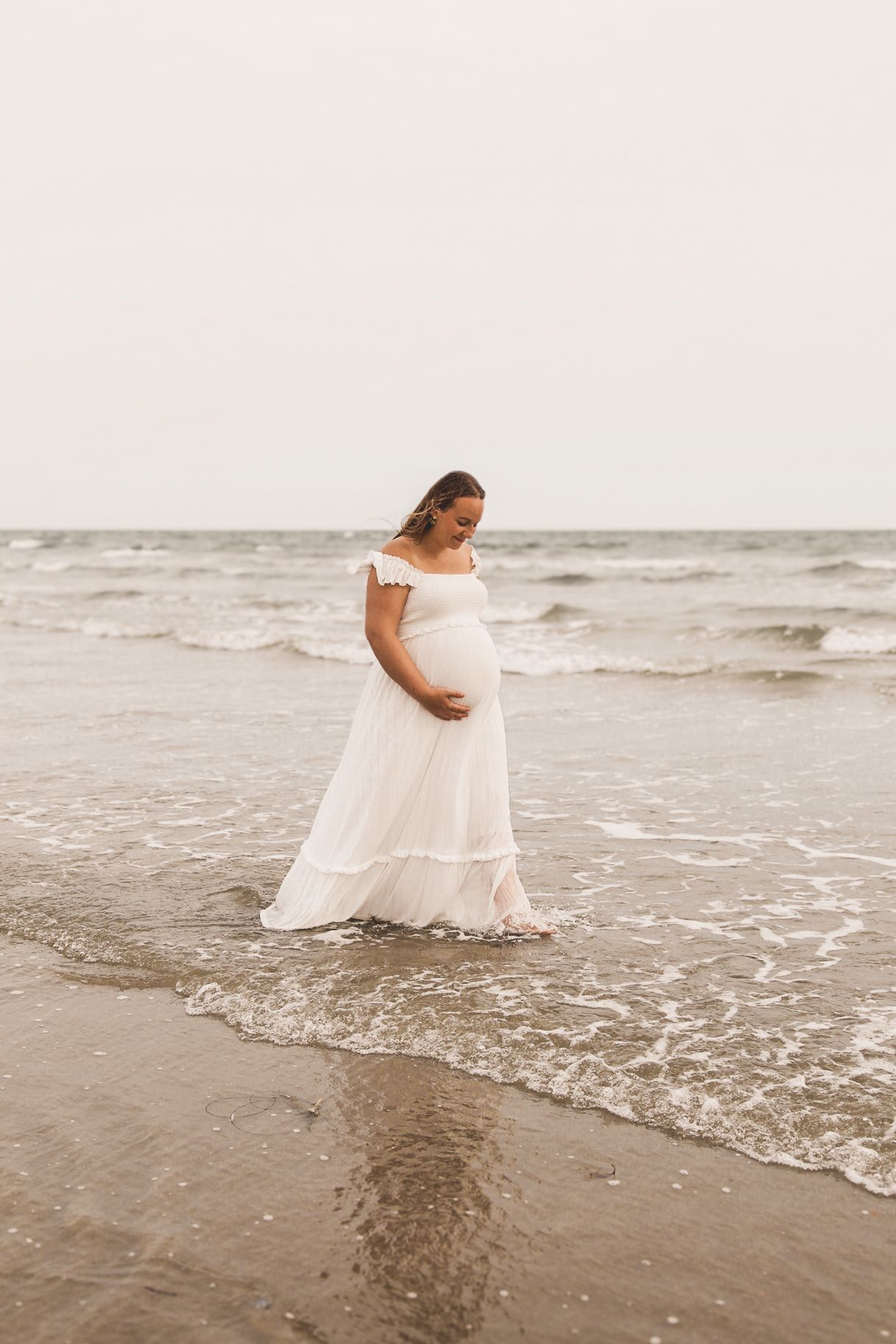 Woman in flowing white dress stands gracefully as waves lap at sandy shore.