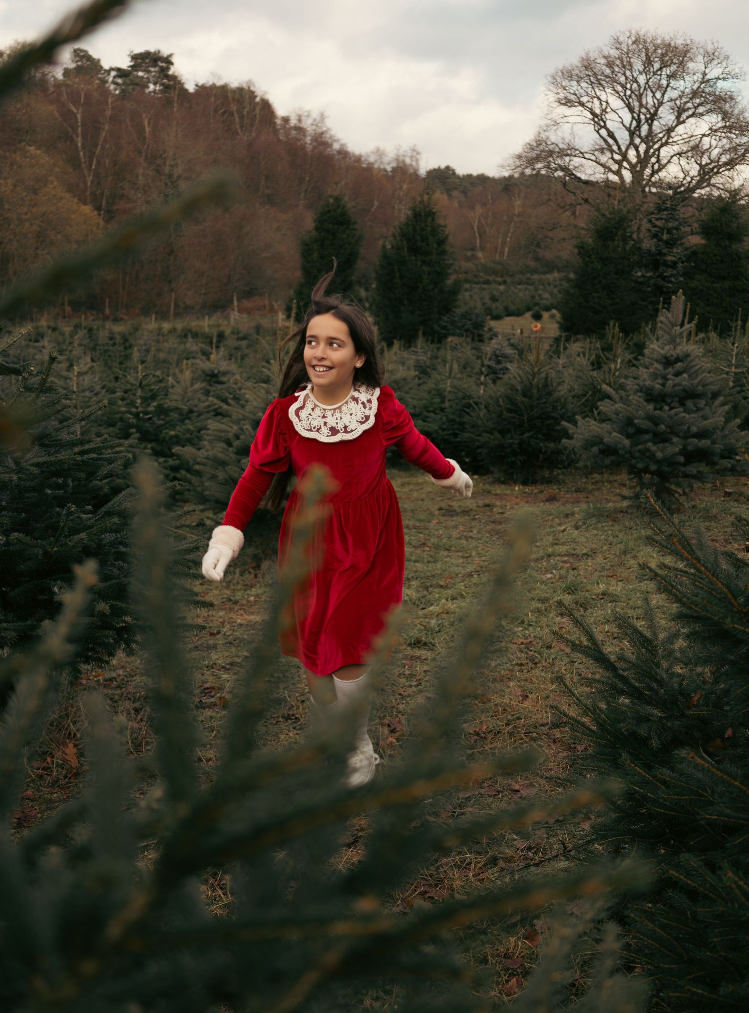 A person in a vintage red dress dances joyfully in a field near evergreen trees.