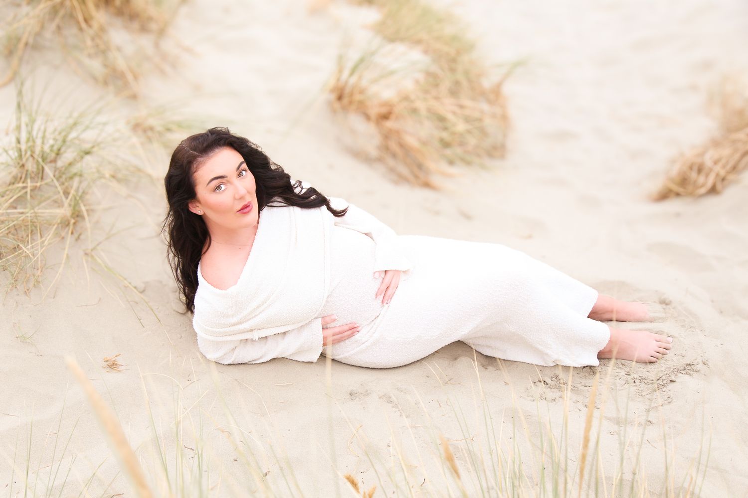 A romantic beach maternity photo with a person in white reclining on sandy dunes.