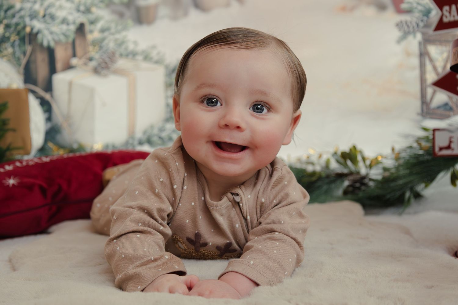 Baby in beige outfit lying on cream carpet with holiday decorations in background.