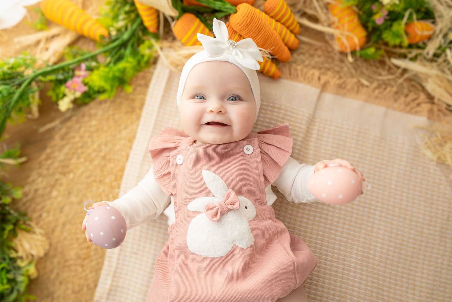 Smiling baby girl in pink Easter outfit with bunny applique and white bow headband, lying among carrots and greenery.