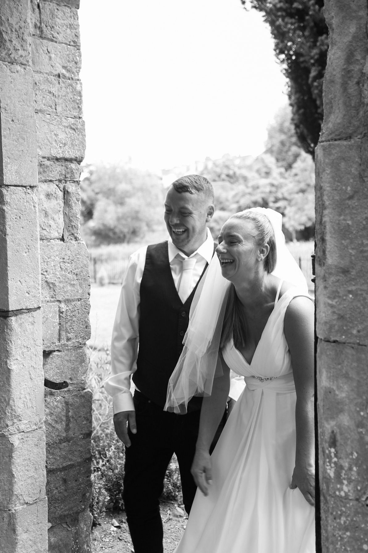 A couple shares a joyful moment between stone pillars in black and white photography.
