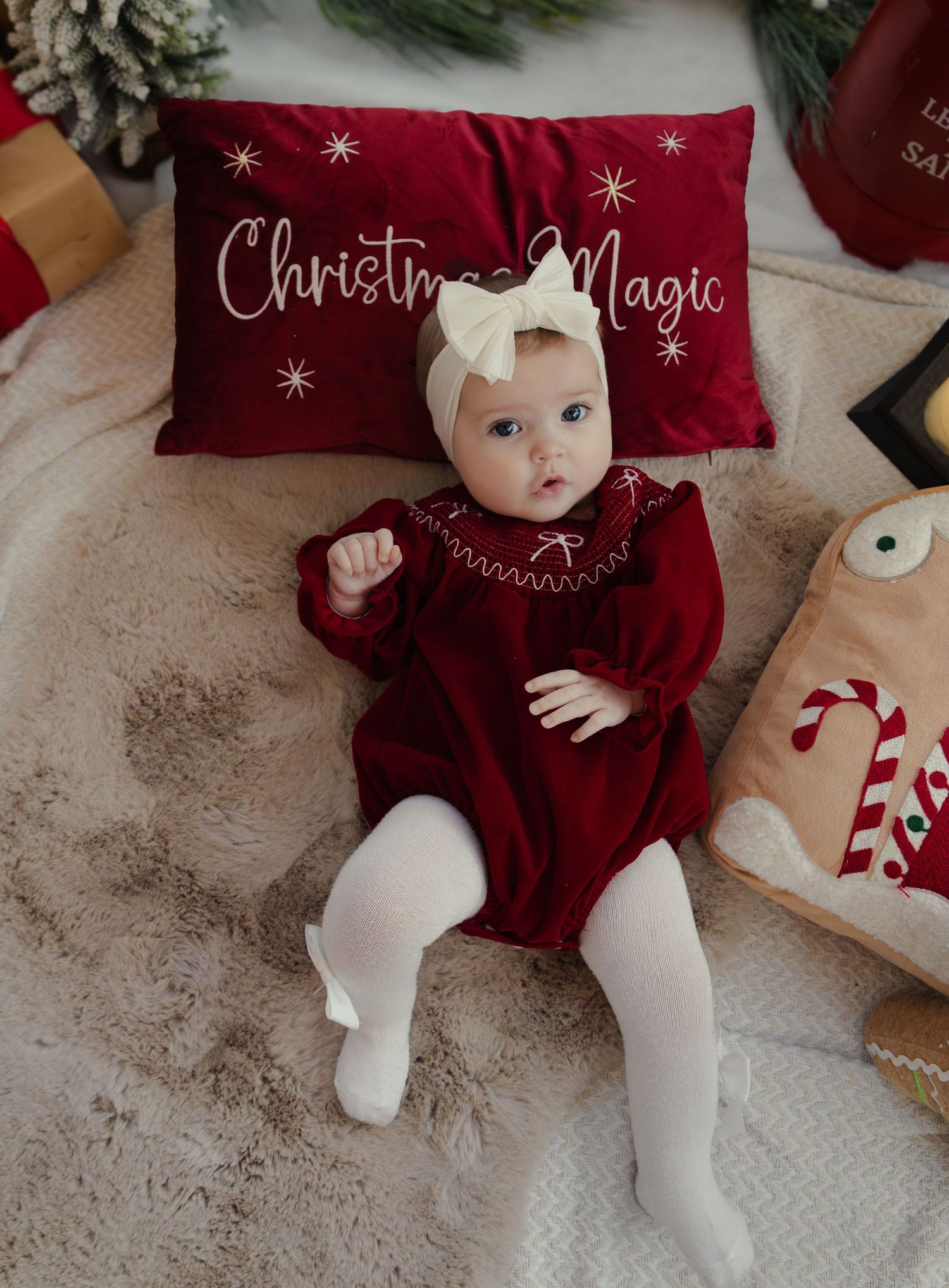 Baby in red velvet dress and white bow lying next to Christmas Magic pillow.