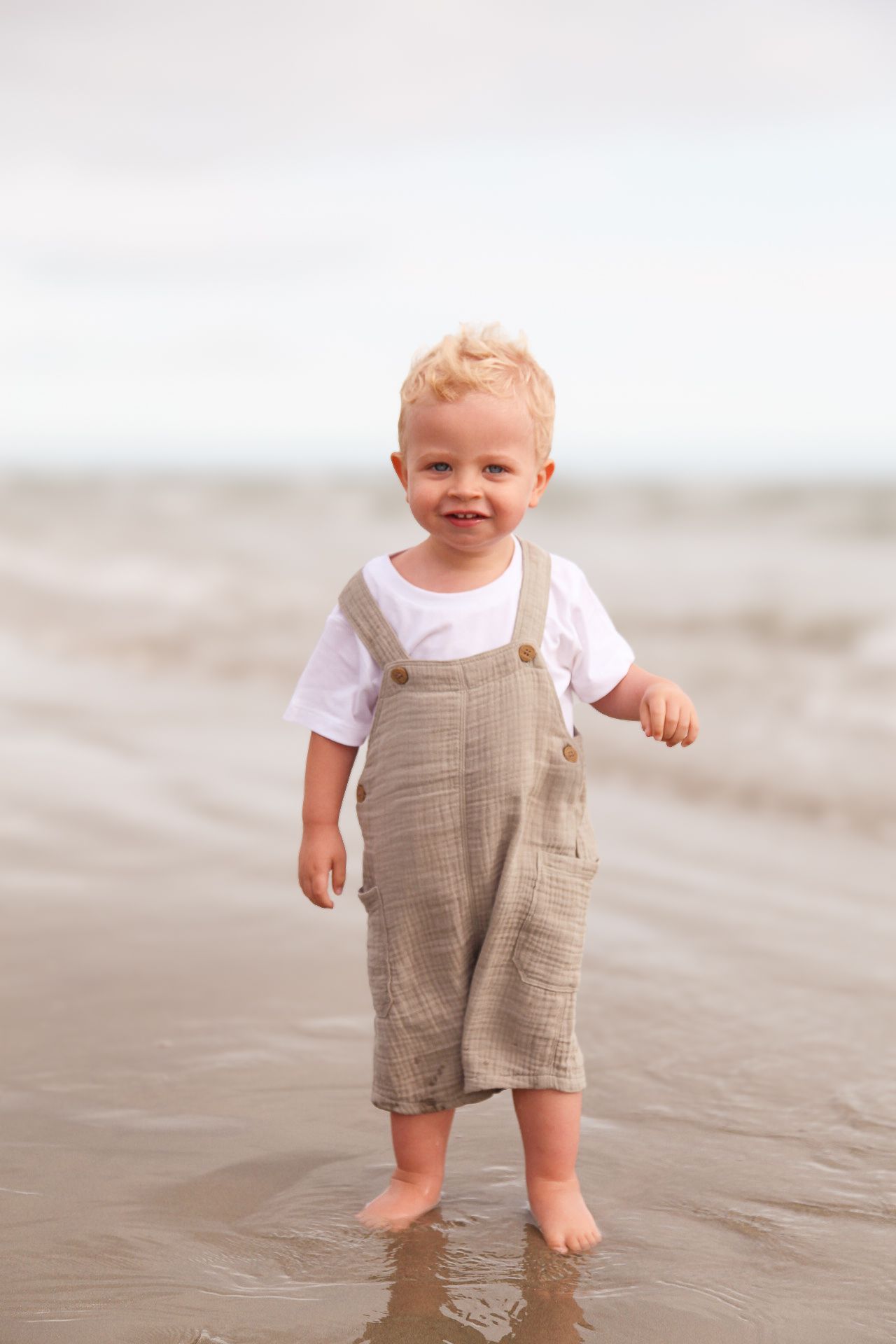 Toddler in beige overalls and white shirt plays on wet beach sand.