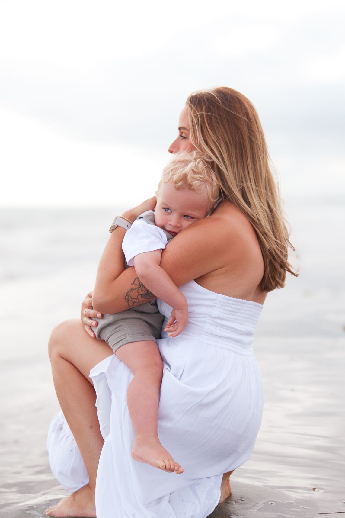 Mother in white dress embracing baby in casual clothes on a peaceful beach setting.