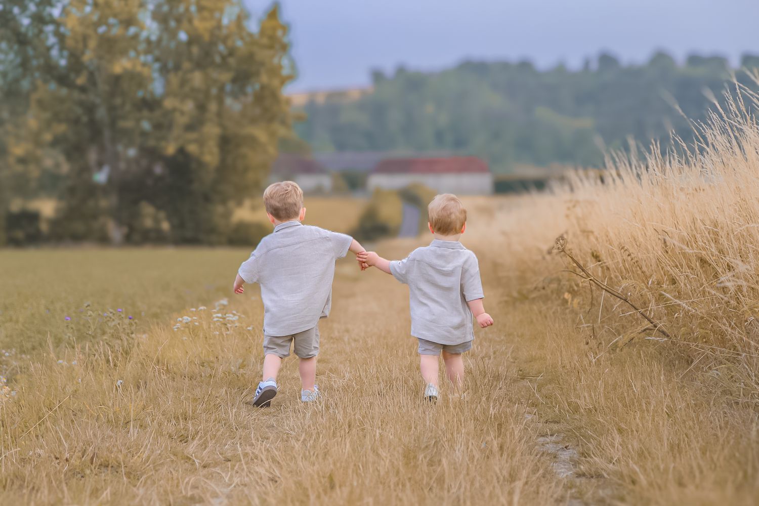 Two small children walk together through a golden wheat field at sunset.