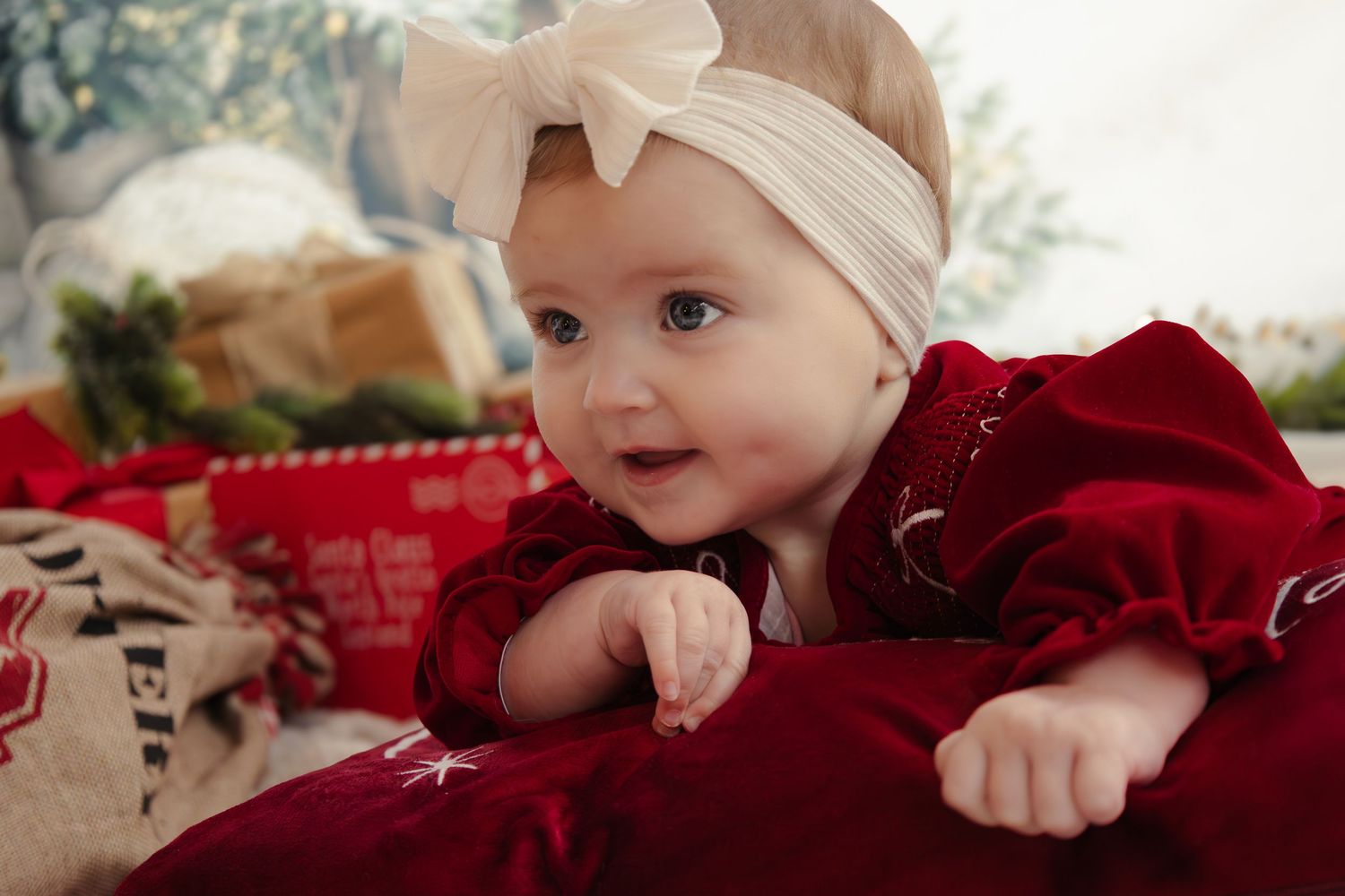Baby in red holiday outfit with white bow headband smiling among Christmas decorations.