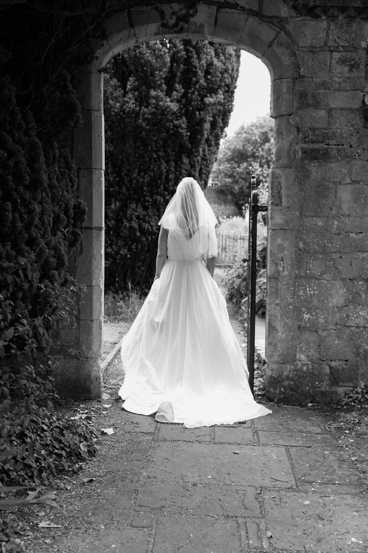A bride in a flowing white dress stands in a stone archway surrounded by ivy.