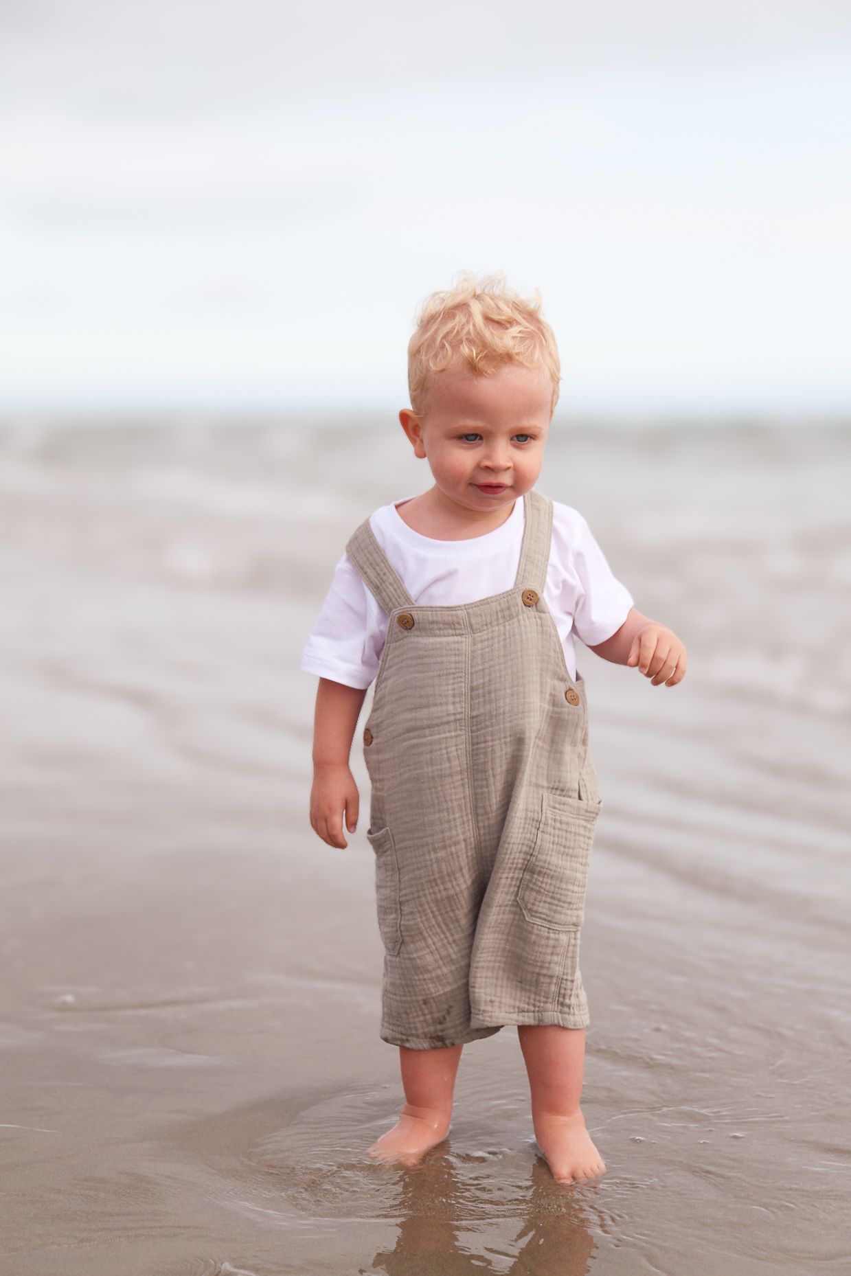 Small child wearing tan overalls stands barefoot in wet beach sand.