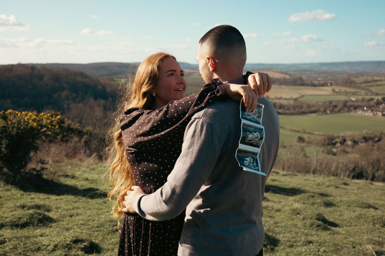 Couple embracing on a scenic hilltop with rolling countryside views on a sunny day.