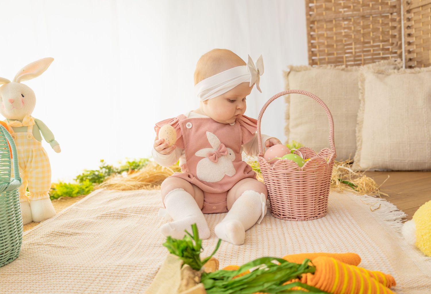 Baby in pink bunny romper sits beside a wicker Easter basket surrounded by carrots and stuffed bunnies.
