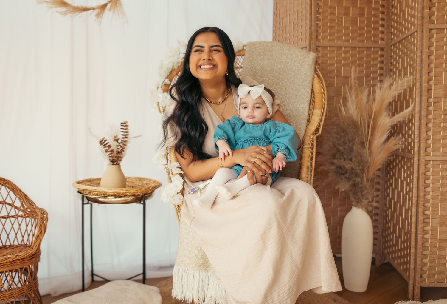 A smiling woman with long dark hair sits in a wicker chair holding a baby in a boho-styled room with neutral decor.