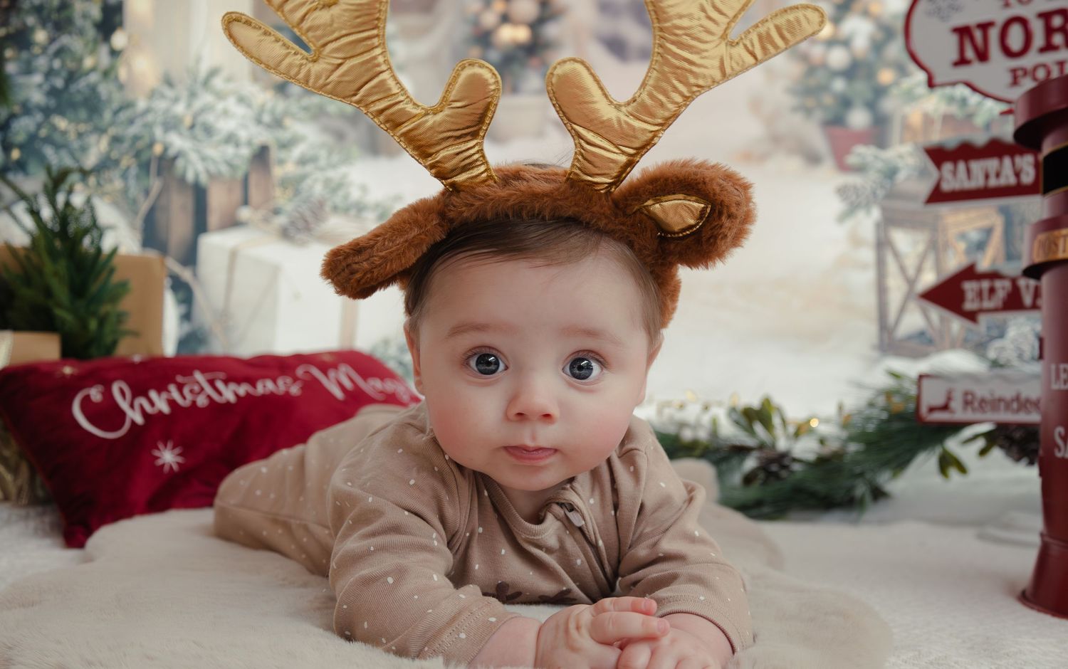 Baby wearing reindeer antler headband and beige outfit lying on floor with Christmas decor.