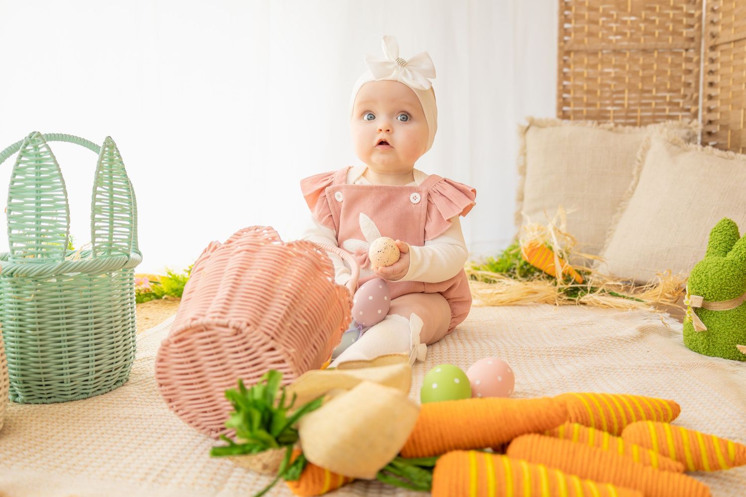 Baby girl in pink romper sits among Easter decorations with colorful eggs, carrots, and wicker baskets.