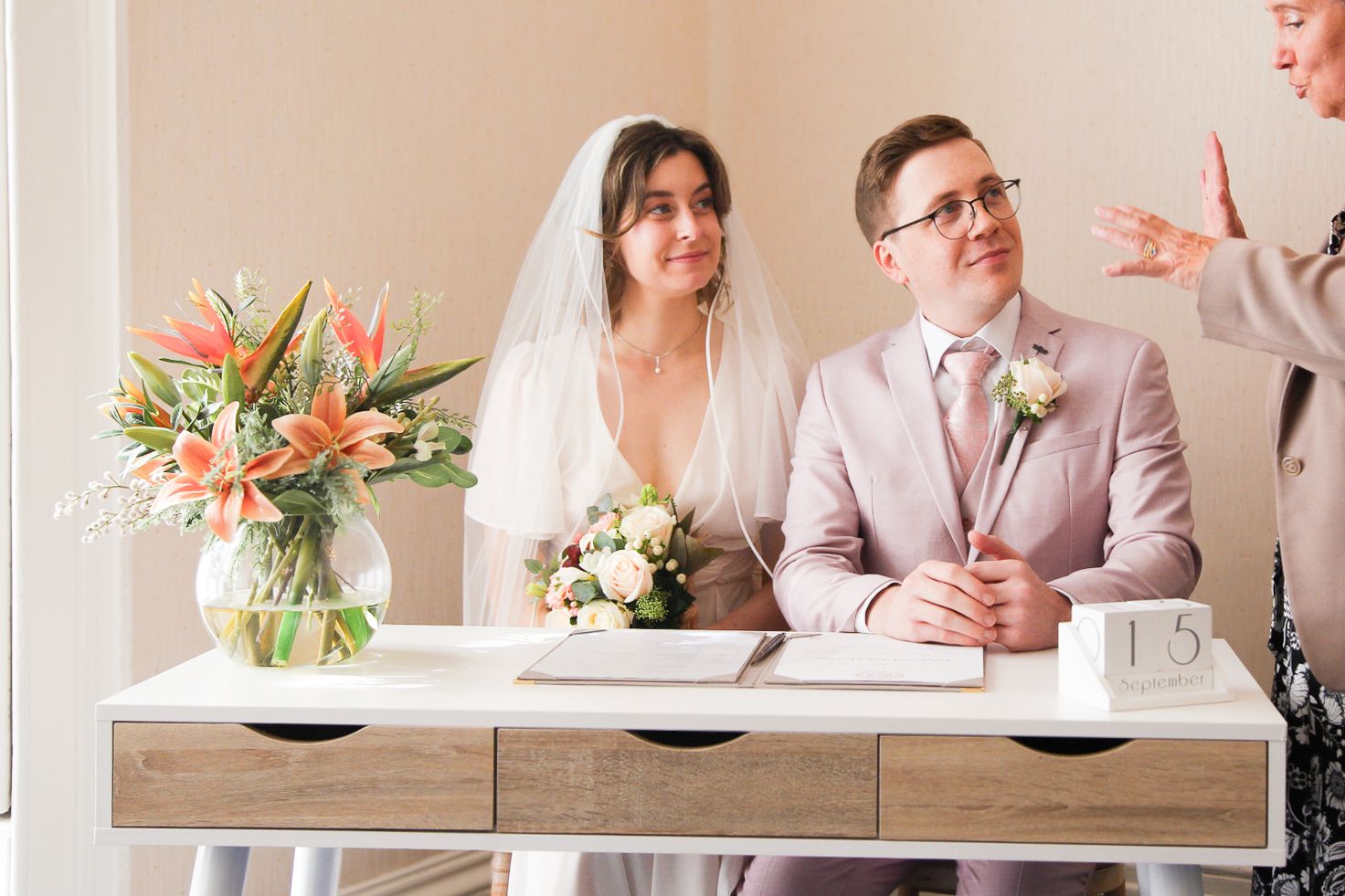 Couple in pink suit and white dress signing marriage documents at elegant venue.