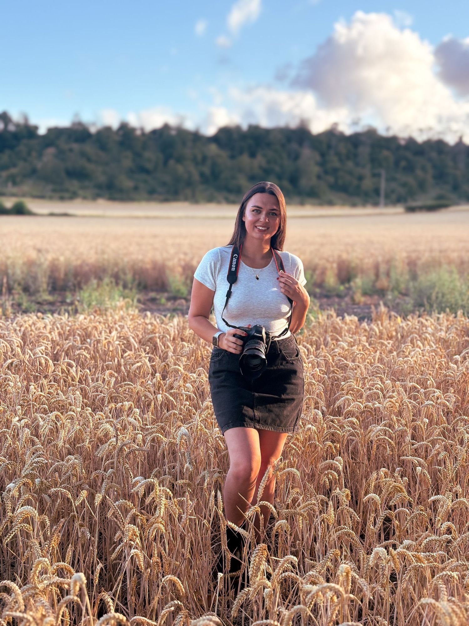 Woman smiling in a golden wheat field holding a camera, wearing a white top and dark skirt on a sunny day.