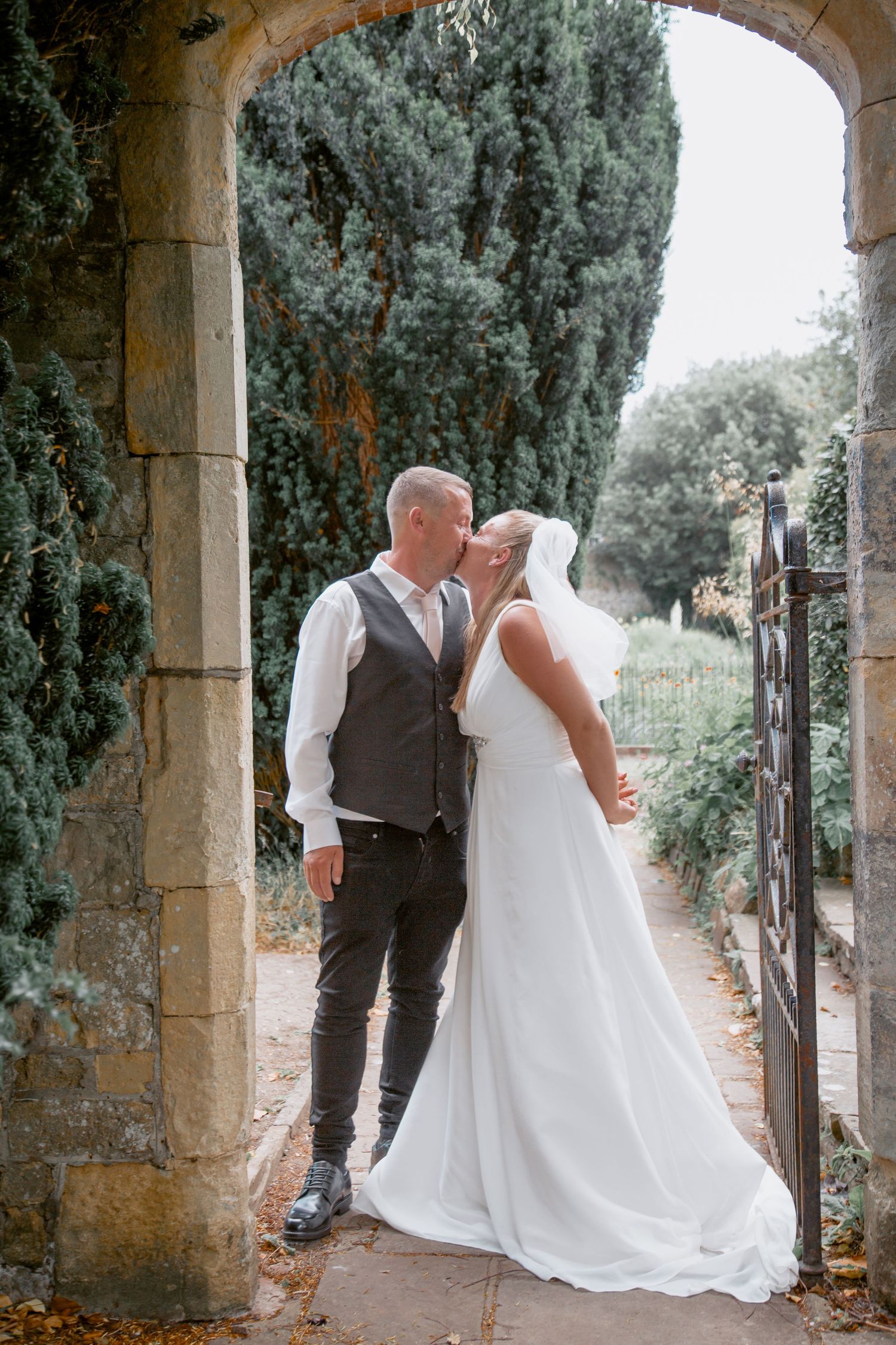 A wedding couple shares an intimate moment under a stone archway with cypress trees in the background.