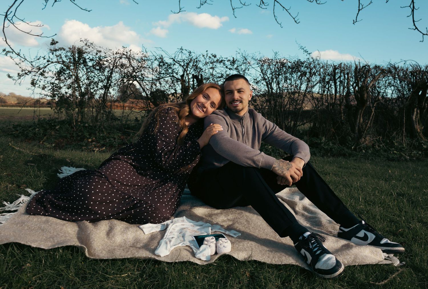 Couple sitting on a blanket in a sunny field, smiling together outdoors near bare trees.