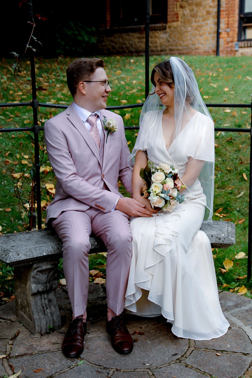 Couple in wedding attire sitting together on stone bench in garden setting.