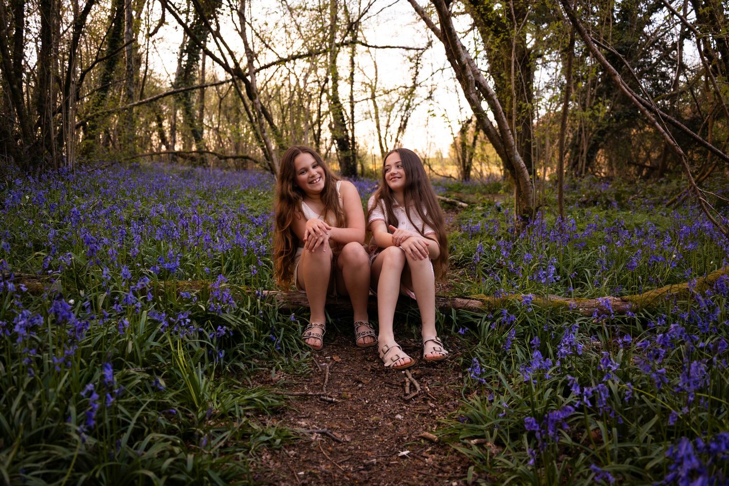 Two girls smiling and sitting on a log surrounded by blooming bluebells in a sunlit woodland forest.