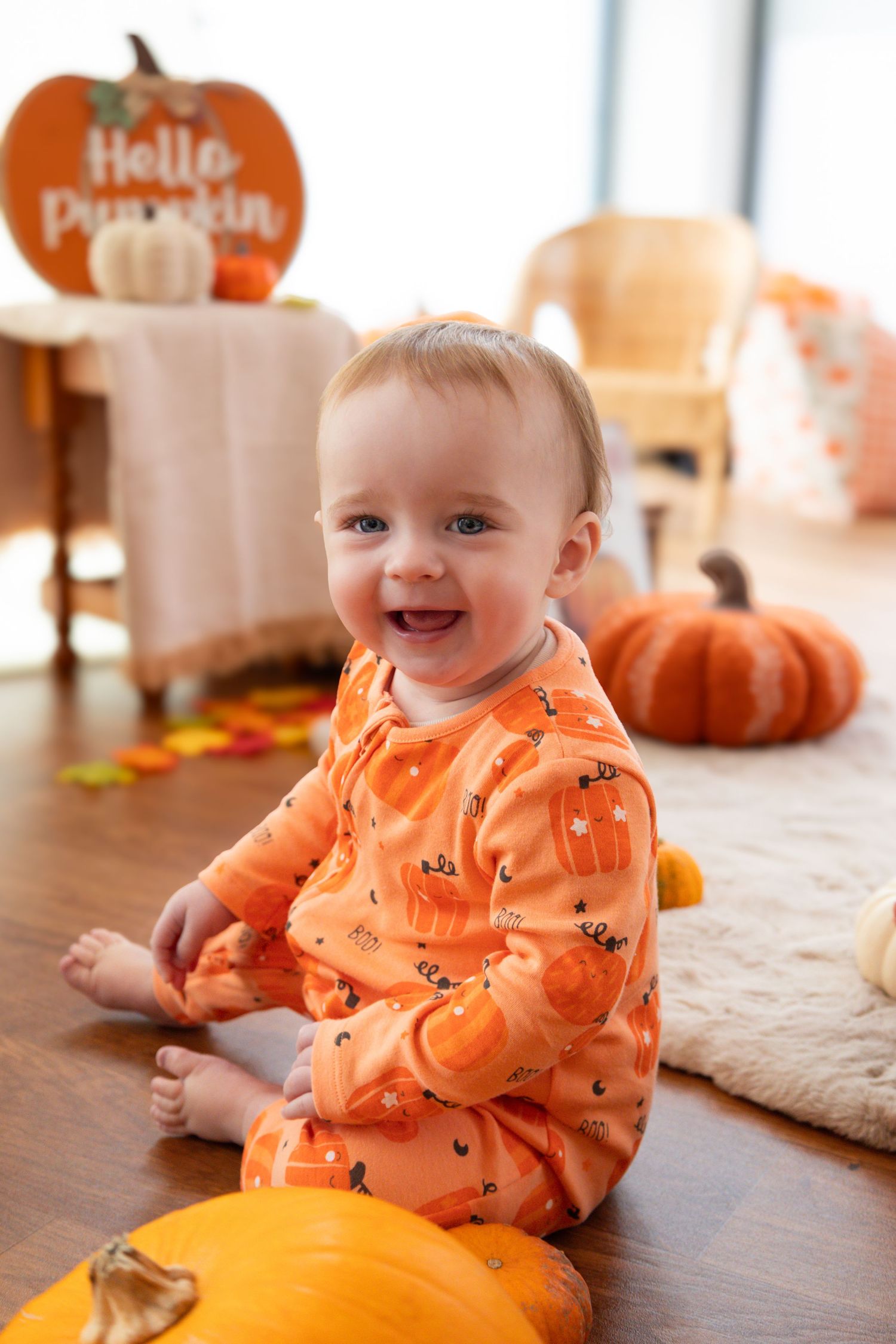 Baby wearing orange Halloween pajamas sitting happily next to pumpkins on hardwood floor.