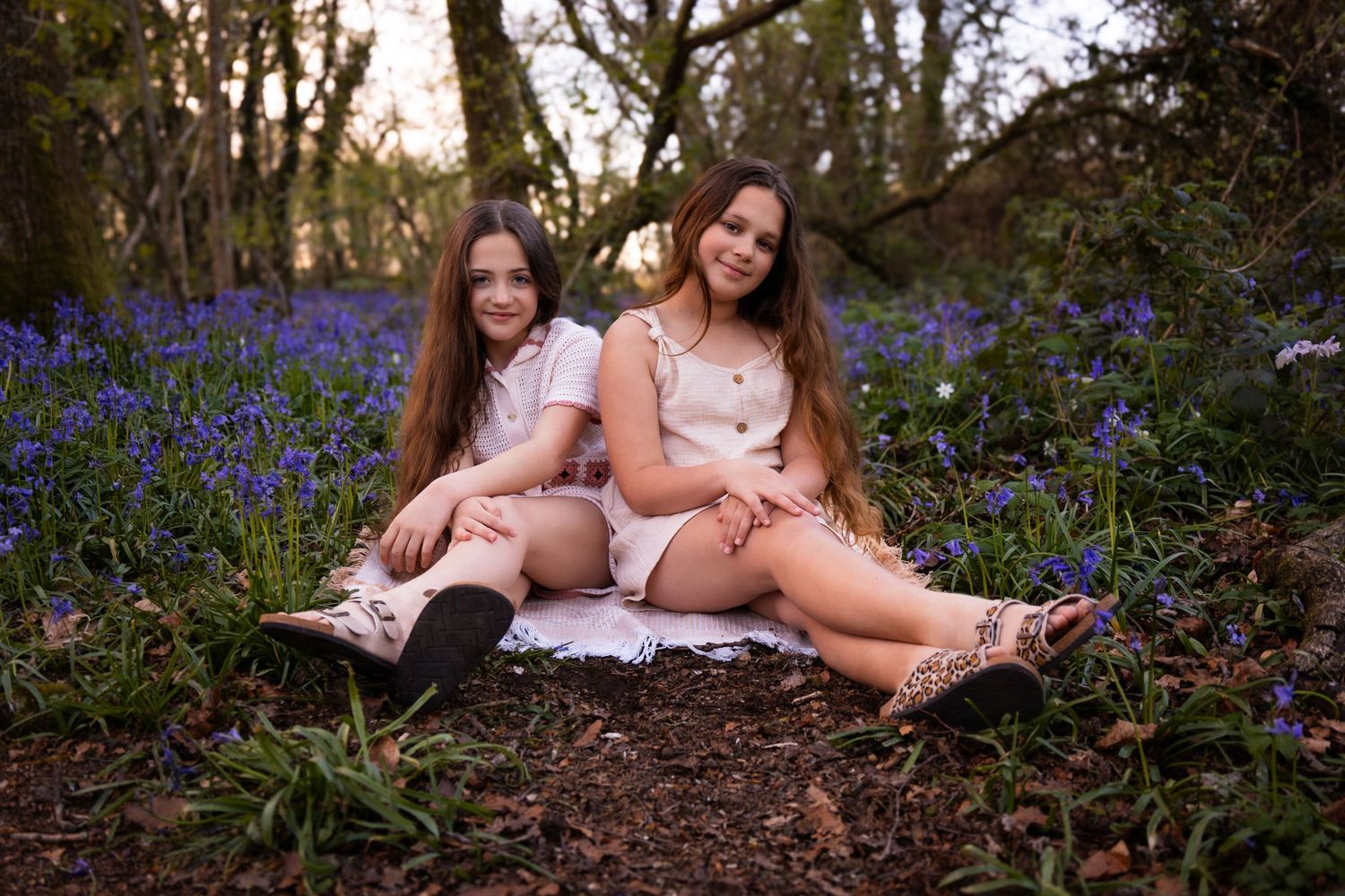 Two girls sitting together in a bluebell woodland, wearing white dresses, surrounded by purple flowers at dusk.