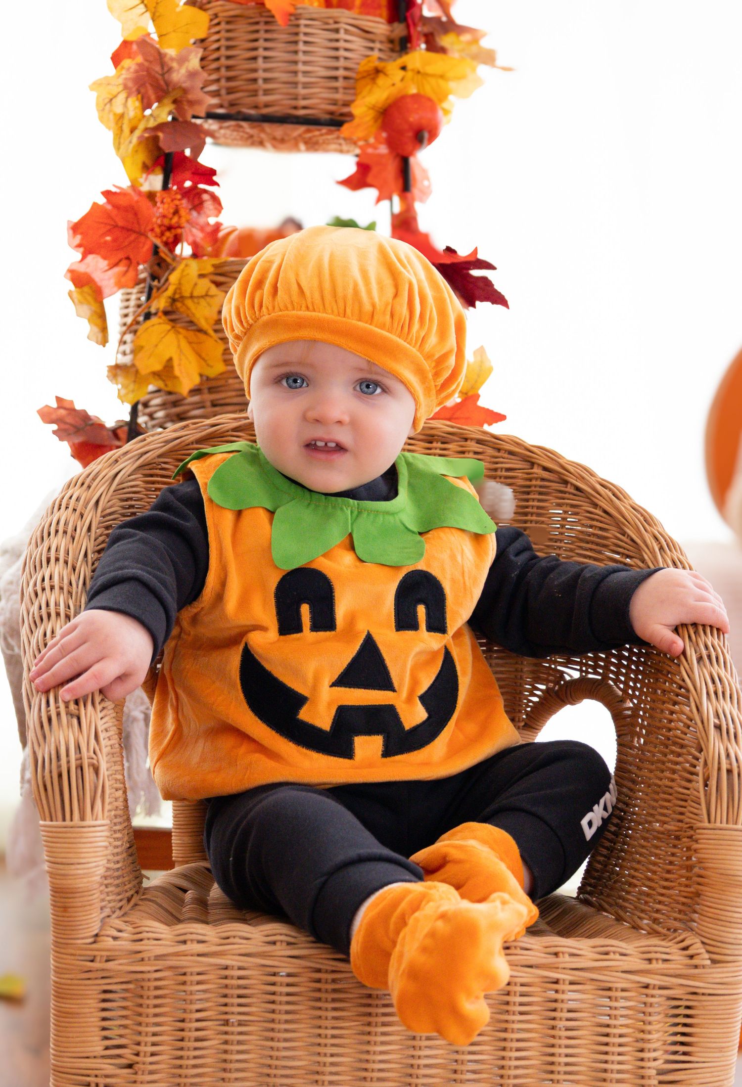 Adorable Halloween pumpkin costume photo series on wicker chair with autumn decorations.