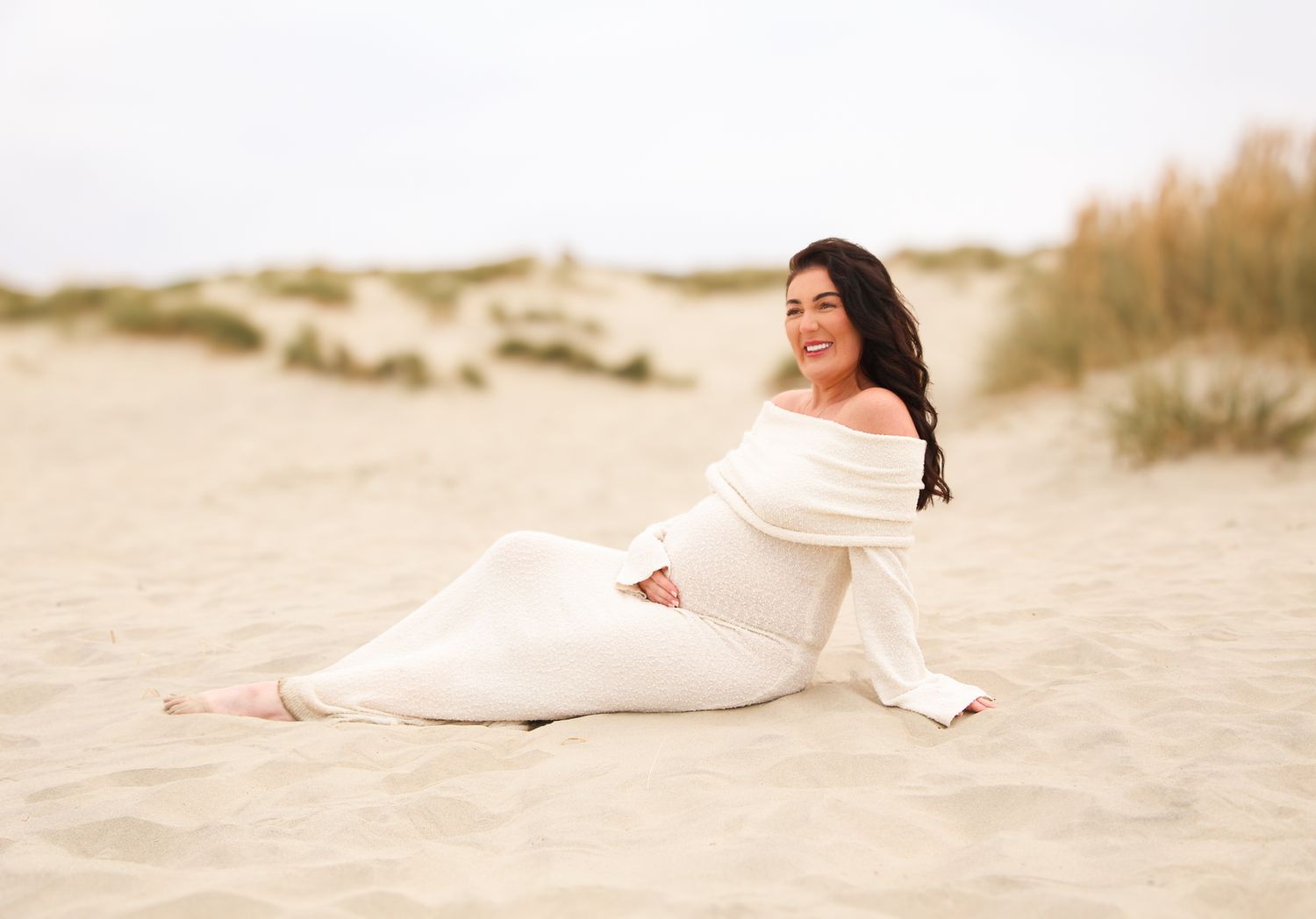 Woman in flowing white dress poses gracefully on sandy beach at sunset.