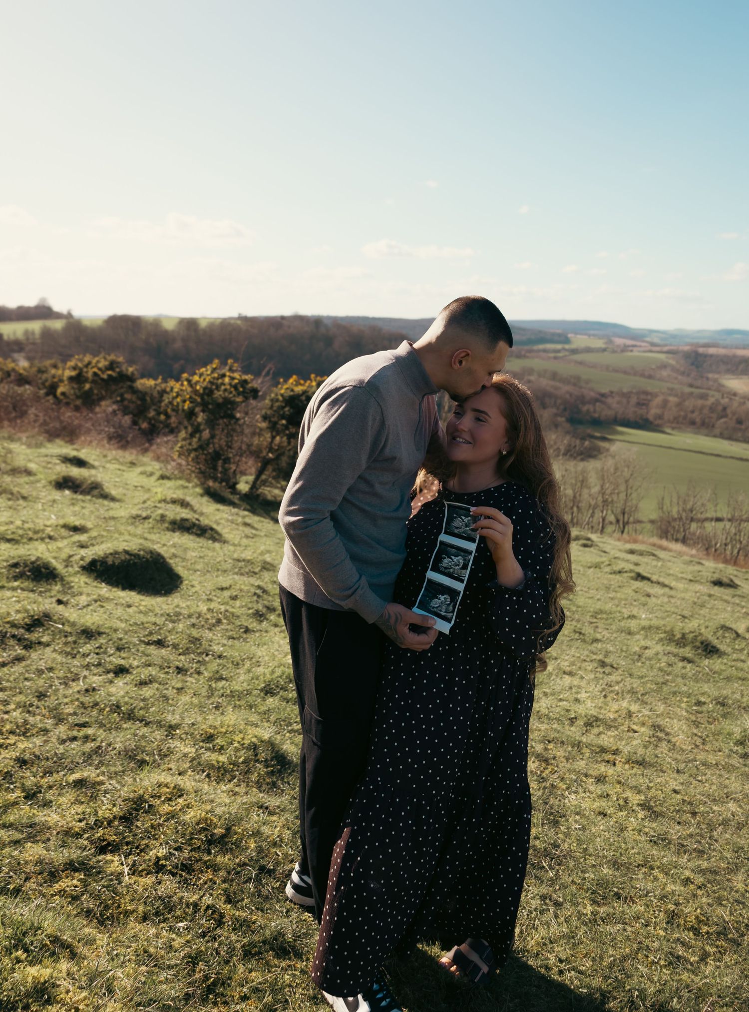 Couple embracing on a green hillside, woman holding ultrasound photos, pregnancy announcement outdoors.