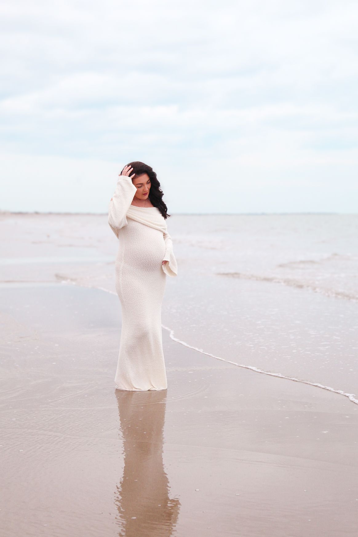 Pregnant woman in white dress standing in shallow ocean waves on a cloudy beach day.